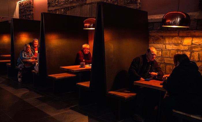 A row of four warmly lit booths; in the first sit a man sits opposite a woman, we see a man in the second and in the third a man and a woman sit next
